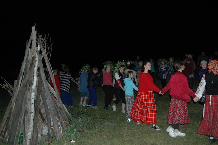 People in flowered wreaths and traditional dress dance around a building fire. 