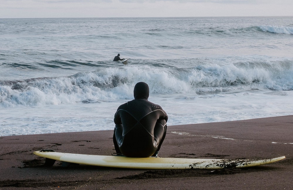 A person in a wetsuit sits on a surfboard and watches their friend surf in the sun. 
